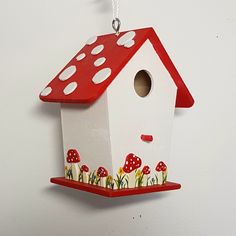 a red and white birdhouse hanging from a chain on a wall with mushrooms around it