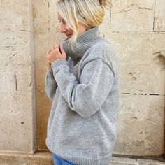 a woman standing in front of a stone wall wearing a gray sweater and blue jeans