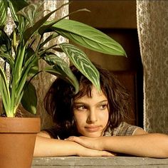 a young woman leaning on a table next to a potted plant with green leaves