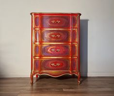 a red and gold painted chest of drawers in a room with hardwood floors, white walls and wood trimmings