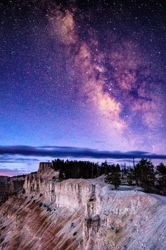 the night sky is filled with stars and clouds above a rocky cliff face, as seen from below