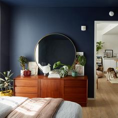 a bedroom with dark blue walls and wooden furniture in the foreground is a large round mirror