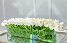 a clear vase with white flowers in it on a mirrored table next to a mirror