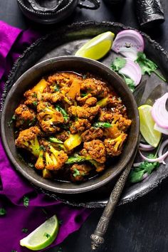 a bowl filled with meat and vegetables on top of a purple cloth next to lime wedges