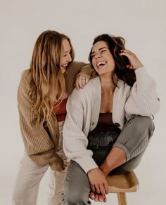 two young women sitting on top of a stool smiling at each other while they both have their arms around one another