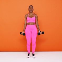 a woman in pink is holding two dumbs and posing for the camera with an orange wall behind her