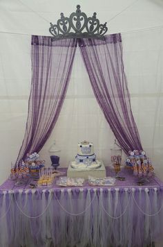 a table topped with a cake covered in purple frosting next to a sheer curtain