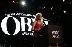 a woman standing at a podium in front of a sign that says the village voice - fly