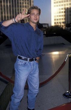 a man standing in front of tall buildings giving the peace sign with his hand while wearing blue jeans and black boots