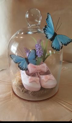 two blue butterflies sitting on top of pink baby shoes under a glass clochet