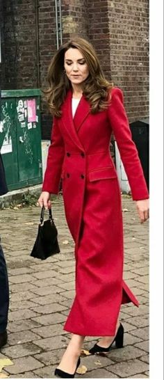 a woman in a red coat and heels walking down the street with a handbag