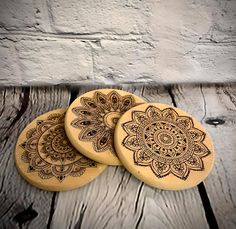 three wooden coasters with intricate designs on them sitting on a wood table next to a brick wall