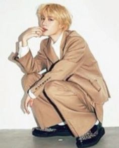 a man sitting on top of a floor next to a white wall wearing a brown coat