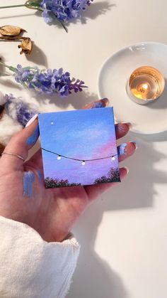 a hand holding a small card with string lights on it and lavender flowers in the background