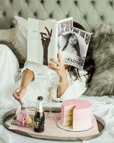 a woman laying in bed reading a magazine and holding a slice of cake next to her face