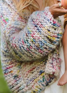 a woman holding an apple in her right hand and wearing a knitted sweater over her left shoulder