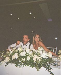 a man and woman sitting at a table with white flowers on the centerpieces