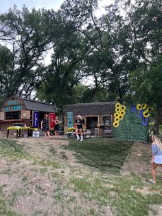 some people are standing in the grass by some food stands and trees with sunflowers on them