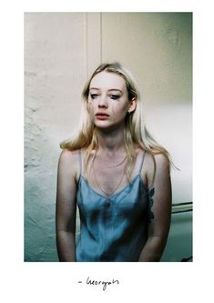 a woman with long blonde hair standing in front of a wall and looking at the camera