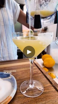 a person pouring a drink into a glass on top of a wooden table with lemons in the background