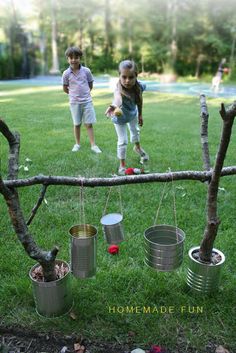 two children playing with tin cans on a tree branch in the grass, and one child is holding an apple