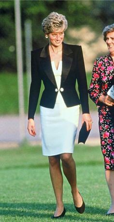 two women are walking in the grass with one woman wearing a black jacket and white dress