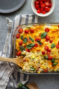 a casserole dish with tomatoes, cheese and spinach on the side next to a wooden spoon