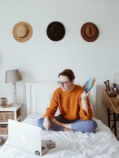 a woman is sitting on her bed while looking at the computer screen and holding an open book