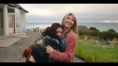 two women hugging each other in front of a house with the ocean in the background