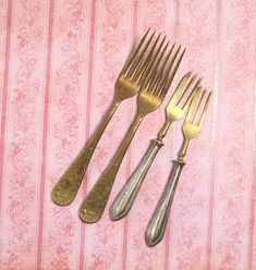 there are three forks and two knives on the pink tablecloth with gold trimmings