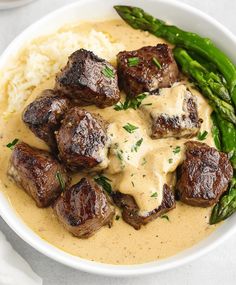 a bowl filled with meat and gravy on top of mashed potatoes next to asparagus