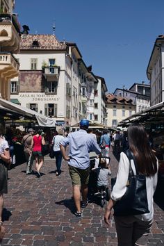 people are walking through an open air market on a cobblestone street with buildings in the background