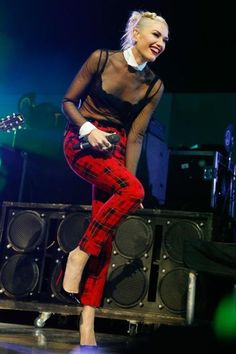 a woman in plaid pants and sheer top on stage with guitar behind her, smiling at the camera