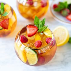 two glasses filled with fruit and ice on top of a white table next to sliced lemons, raspberries, and mint