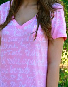 a woman wearing a pink shirt with white writing on the front and back, standing in front of some trees