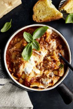 a bowl of pasta with meat and basil on top, next to slices of bread