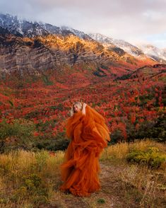 a woman in an orange dress standing on top of a grass covered field next to mountains