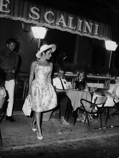 black and white photograph of people sitting at tables in front of a restaurant with lights on