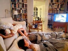 two people are laying on the couch watching tv in a living room with bookshelves