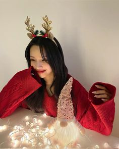 a woman wearing a red sweater with reindeer antlers on her head, sitting in front of christmas lights