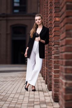 a woman leaning against a brick wall wearing white pants and a black blazer jacket