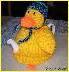 a knitted yellow duck sitting on top of a bed next to a tea pot