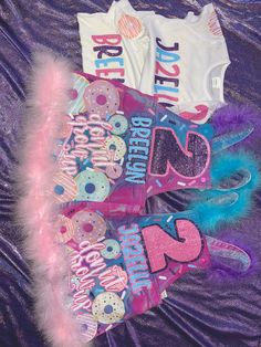 three birthday shirts are laying on a purple sheet with pink and blue feathers around them