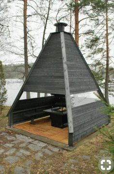 a small black cabin in the woods with a deck and picnic table under it's roof