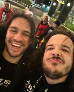 two men are smiling for the camera while standing on a sidewalk at night with other people in the background