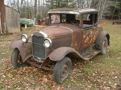 an old rusted out car is parked in the grass next to some trees and leaves