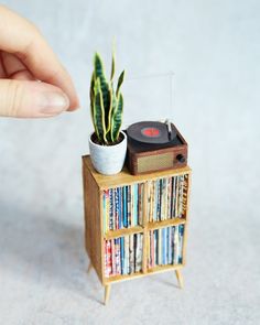 a hand holding a record player next to a wooden shelf filled with cds and a plant