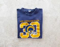 "70's L.A. Rams t-shirt in good vintage condition. Size tag marked size L but would fit modern day women's size small best. Please refer to all photos for wear. Length: 23.5\", shoulder to hem Width: 17\", pit to pit (Measured flat) A note on vintage t-shirt sizing: To ensure a perfect fit, we recommend measuring your favorite t-shirt and comparing it to the measurements specified above for this item. PLEASE READ BEFORE PURCHASE: All of our items are pre-loved vintage and may show signs of wear. Vintage Blue Tops For Fan Merchandise, Blue Retro T-shirt For Sports Events, Retro Blue T-shirt For Sports Events, La Rams Football, Rams Football, Football Game Outfit, La Rams, Gaming Clothes, Vintage Tshirts