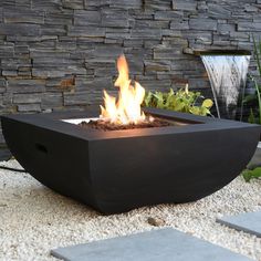 a fire pit sitting on top of a gravel field next to a stone wall and water fountain