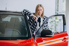 a woman leaning on the hood of a red car and pointing at something in her hand